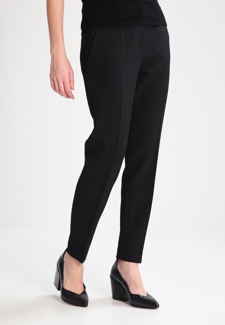 Femme portant un pantalon de costume noir fuselé et des chaussures à talons carrés noires à bout fermé, debout devant un fond blanc uni.