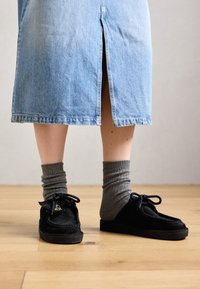 Personne portant des mocassins en suède noirs avec des chaussettes grises côtelées et une jupe en denim bleu clair avec une fente à l'arrière, se tenant sur un sol en bois.