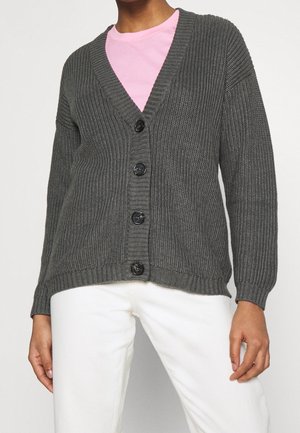 Personne portant un cardigan gris côtelé boutonné sur une chemise rose, associé à un pantalon blanc, debout avec les bras détendus.
