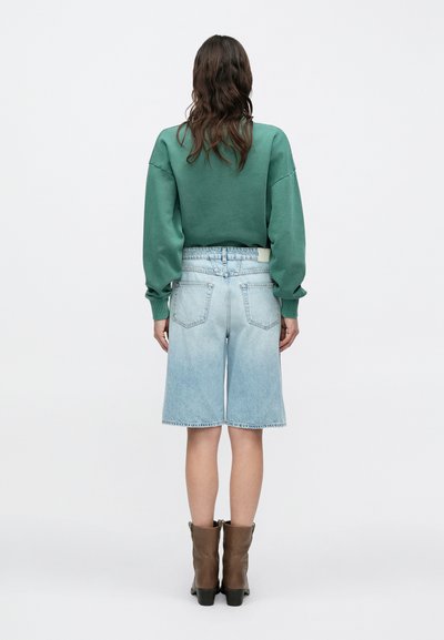 Κοντό πράσινο φούτερ με κατεβασμένους ώμους, συνδυασμένο με ανοιχτόχρωμα μπλε τζιν σορτς, με ξεθωριασμένο πλύσιμο και ορατές πίσω τσέπες, φορεμένο με καφέ μποτάκια.