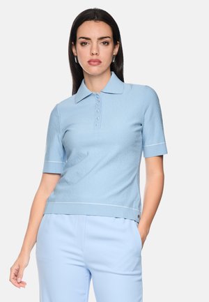 Frau trägt ein hellblaues, kurzärmeliges Strickshirt mit Kragen und Knöpfen sowie hellblaue Hose, steht mit einer Hand in der Tasche.