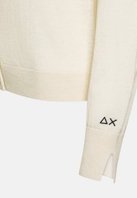 Cardigan lavorato a maglia di colore crema con una texture a coste, caratterizzato da un dettaglio a spacco sui polsini e un logo geometrico nero ricamato.