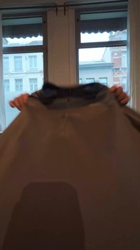 Persona tur brūnu kapuci telpās ar lieliem logiem, caur kuriem redzamas ēkas fona.