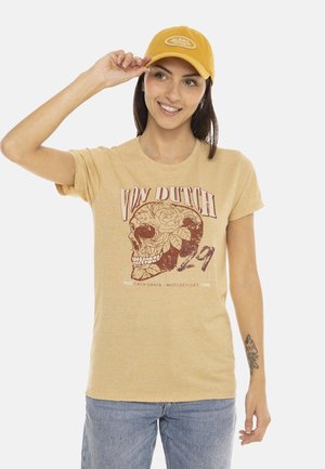 Femme portant un t-shirt beige Von Dutch avec un motif de crâne et une casquette jaune, souriante et touchant la visière de la casquette.