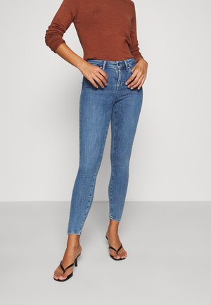 Femme portant un jean skinny bleu taille haute, un haut à manches longues couleur rouille et des sandales noires à talons sur un fond uni.
