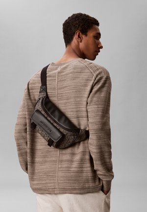 Hombre con suéter beige a rayas y bolso cruzado con patrón y detalles de cuero negro, de pie frente a un fondo gris, mirando de lado.