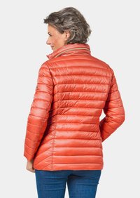 Orangene gepolsterte Jacke mit hohem Kragen, Reißverschluss und genähten horizontalen Linien. Über blauen Jeans getragen. Glatte, glänzende Textur.
