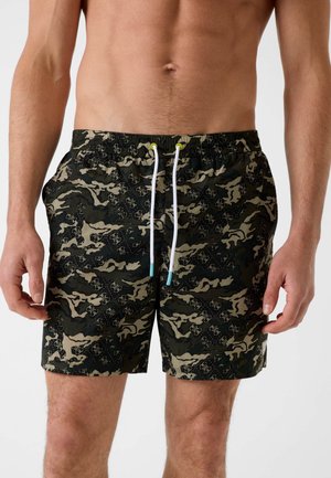 Herrenshorts mit Tarnmuster, weißen Kordeln und hellblauen Enden, getragen von einem oberkörperfreien männlichen Model vor einem einfarbigen Hintergrund.