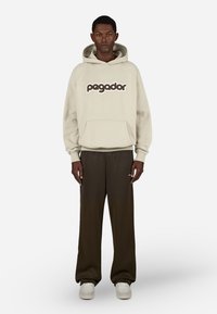 Beiger Hoodie mit dem "pagador"-Logo in Dunkelbraun, kombiniert mit lockeren, dunkelgrünen Hosen. Der Stoff wirkt weich mit einer geräumigen Fronttasche.