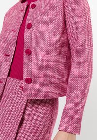 Veste en tweed rose avec une coupe carrée, deux poches avant et quatre boutons ronds. Tissu texturé avec un détail intérieur contrastant.