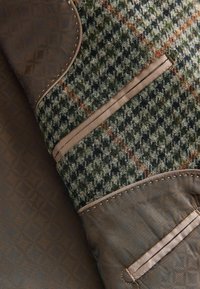 Uldblandingsstof i et grønt, sort og orange hundetand-mønster. Læderdetaljer i brun med dekorativ syning og lommer.