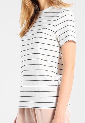 Vrouw die een wit shirt met korte mouwen draagt met dunne zwarte horizontale strepen en lichtroze broek, getoond van schouders tot heupen.