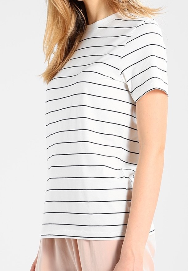 Femme portant une chemise blanche à manches courtes avec de fines rayures horizontales noires et un pantalon rose clair, montrée des épaules aux hanches.