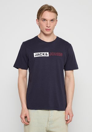 Jonge man die een marineblauw T-shirt draagt met de tekst "JACK & JONES" in wit en rood, gecombineerd met lichtbeige broek, staand tegen een effen achtergrond.