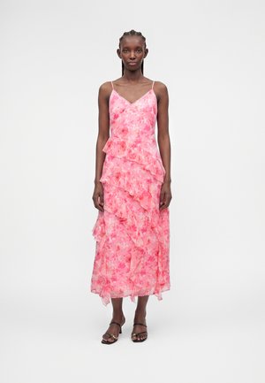 Γυναίκα με πλεξούδες στα μαλλιά, που φοράει ροζ φόρεμα με floral μοτίβο, αμάνικο, μήκους μέσης με βολάν και μαύρα ψηλοτάκουνα σανδάλια με ανοιχτά δάχτυλα, στέκεται μπροστά σε λευκό φόντο.