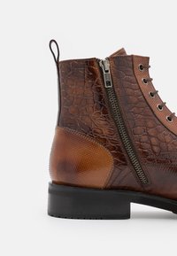 Hnědé kožené kotníkové boty s texturovaným vzorem, bočním zipem, šněrováním a černou gumovou podrážkou na bílém pozadí.