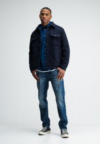 Navy blue wool jacket with two front pockets over a denim shirt, paired with light blue jeans and black sneakers with white soles.