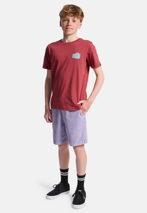Roodharige jongen staat met één hand in zijn zak, draagt een rood t-shirt met print, lichtpaarse korte broek, zwarte sokken en zwarte sneakers.