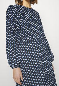 Svart klänning med ett blått och vitt geometriskt mönster, långa ärmar och rynkade detaljer vid halsringningen och ärmsluten, tillverkad av lättviktsmaterial.