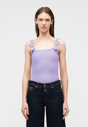 Ženska z dolgimi rjavimi lasmi, oblečena v vijoličasto zgornji del brez rokavov z zaponkami na naramnicah in temne hlače z visokim pasom, stoji pred enobarvno ozadje.