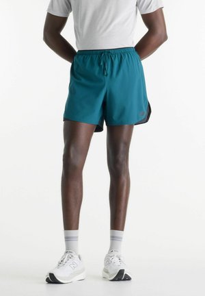 Persona che indossa pantaloncini sportivi verde acqua, calzini bianchi con strisce grigie, scarpe da corsa bianche e una maglietta bianca, in piedi con le mani dietro la schiena.