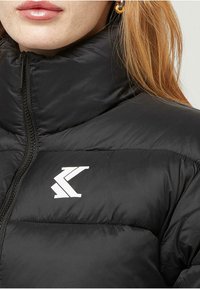 Detailný záber osoby v čiernej prešívanej bunde s vysokým golierom a bielym logom na hrudi, pričom je vidieť časť jej tváre a vlasov.