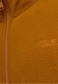 Veste en polaire jaune moutarde, avec une surface texturée, une fermeture éclair frontale et un logo "Jack Wolfskin" brodé de la même couleur.