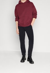 Sudadera con capucha color burdeos con un bolsillo frontal, combinada con pantalones de pana negra de corte ajustado y botas negras de cordones. Textura suave, estilo casual.