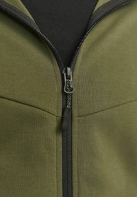 Veste zippée vert olive avec une fermeture éclair noire et une texture lisse, avec un tissu noir contrastant sous le col.