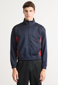 Ναυτική αθλητική ζακέτα με κόκκινες πλάγιες ρίγες, ψηλό γιακά, πλήρη φερμουάρ, ελαστικούς μανσέτες και ελαστική μέση; κατασκευασμένη από λείο, ελαφρύ ύφασ�μα.