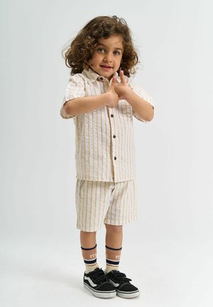 Pruhovaná svetlá béžová a biela košeľa s krátkym rukávom a zodpovedajúce šortky, skombinované s čiernymi teniskami a vzorovanými ponožkami. Jednoduché pozadie.