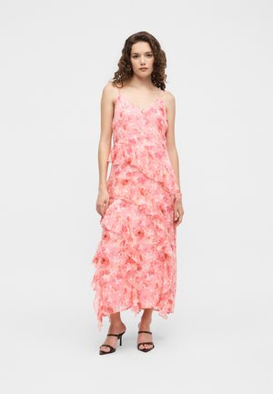 Vrouw met krullend haar die een roze bloemenmouwloze jurk met ruches en zwarte open teen sandalen met hak draagt, staand tegen een effen achtergrond.