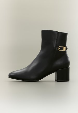 Schwarzer Leder-Stiefeletten mit Blockabsatz und goldenem Schnallendetail an der Seite vor neutralem Hintergrund.