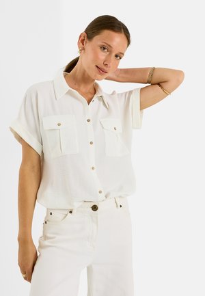 Mujer con cabello castaño en una coleta baja lleva una camisa blanca suelta de manga corta con botones y pantalones blancos, con pendientes y pulseras doradas.