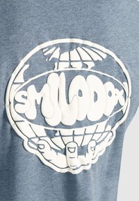 Blåt heathered stof prydes af et stort hvidt broderet grafisk motiv af hænder, der holder en globe, med ordet "SMILADA" i en bølget skrift.