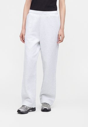 Personne portant un pantalon de jogging ample gris clair, un haut noir et des chaussures de course argentées, debout sur un fond clair uni.