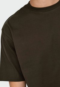 Nahaufnahme einer Person, die ein dunkelolivgrünes Rundhals-T-Shirt mit kurzen Ärmeln vor einem einfarbigen Hintergrund trägt.