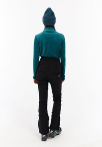 Turkusowa koszula z długim rękawem o fakturowym wzorze, zestawiona z czarnymi spodniami. Czarne buty narciarskie z niebieskimi akcentami. Okrągła wełniana czapka na głowie.