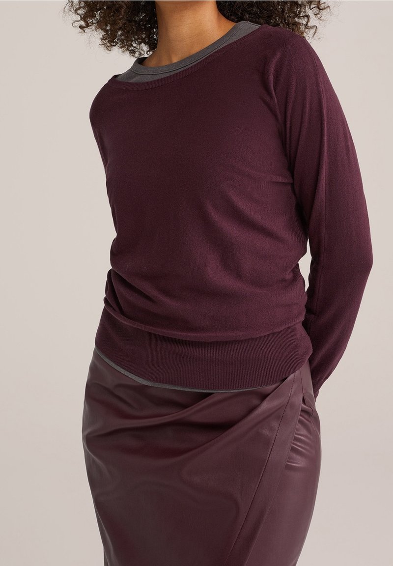Femme portant un pull violet foncé superposé à une chemise grise et une jupe en cuir violet foncé assortie, sur un fond uni.