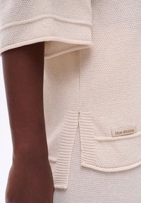 Donkere huidarm naast een crèmekleurige gebreide trui met een geribbelde zoom en een klein "blauw schaduw" label vlakbij de zijsplit.