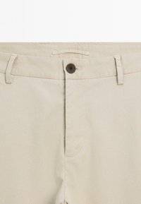 Beige pants waistband with brown button, belt loops, and zipper fly, showing detailed fabric texture.