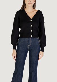 Cardigan nero a coste con scollo a V, dotato di sette bottoni rotondi simili a perle sul davanti e maniche a palloncino. Indossato con jeans blu scuro.