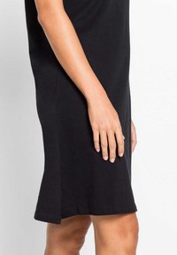 Abito di cotone nero con una vestibilità rilassata, dotato di maniche corte, fessure laterali e una texture liscia. Mano appoggiata sul lato dell'abito.