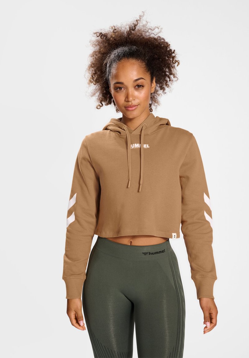 Ung kvinde med krøllet hår iført tan crop-top hættetrøje og olivengrønne leggings med høj talje, stående mod en ensfarvet hvid baggrund.