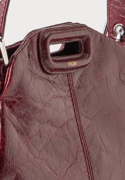 Sac à main en cuir rouge foncé avec poignée découpée rectangulaire et logo doré "moje" sous l'ouverture de la poignée.