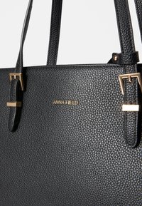 Bolso negro texturizado con dos asas resistentes, acentos de hardware en tono dorado y un sutil logo de la marca. Diseño estructurado con un detalle mínimo.