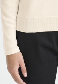 Svetlo krémový pletený sveter s rebrovaným lemom a manžetami, v kombinácii s čiernymi nohavicami, ktorý predstavuje hladkú textúru a pohodlný strih.