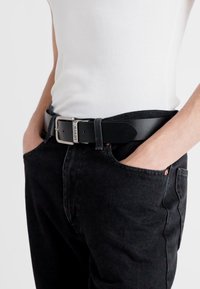 Zwarte leren riem met een gestructureerd oppervlak, zilveren metalen gesp gegraveerd met "Levi's," gedragen over donkere spijkerbroeken en een witte tanktop.