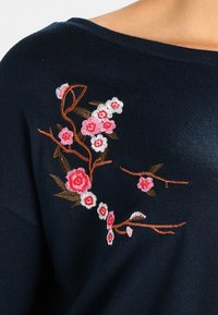 Marineblauwe stof met geborduurde roze en witte kersenbloesems op bruine takken nabij de schouder.