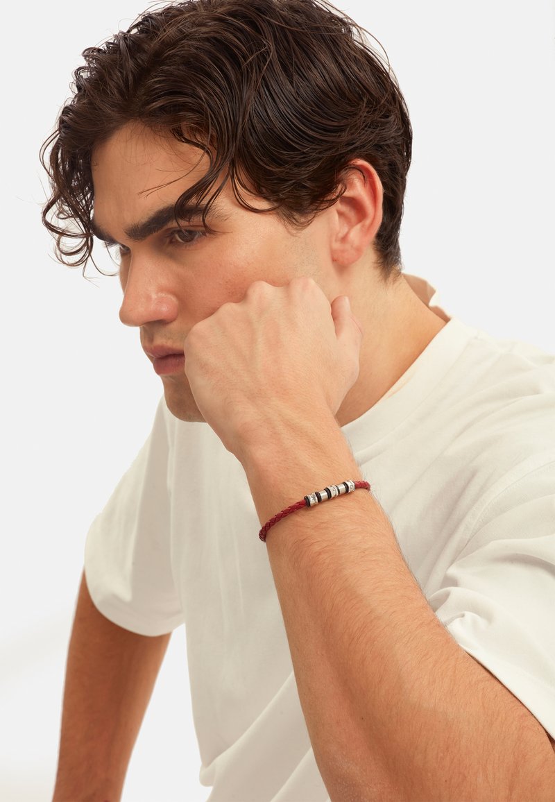 Pulsera trenzada roja con acentos cilíndricos metálicos, llevada en una muñeca, sobre un fondo blanco liso.